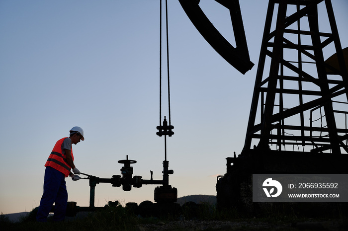 Petroleum operator in work vest using oil pump rocker-machine to extract crude oil from well. Male worker operating petroleum pump jack while working in oil field. Concept of oil extraction.