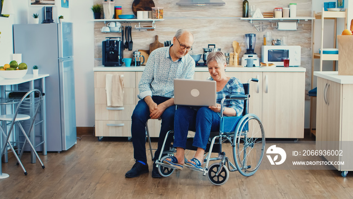 Senior couple waving at webcam during a video call on laptop in kitchen. Paralysied handicapped old elderly woman and her husband on online call, using modern communication tech.