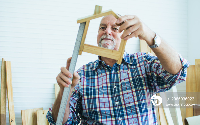 Senior Caucasian happy retired old male woodworker or carpenter smiling with happiness, wearing check shirt, measure to create DIY wooden home model for house decoration. Hobby, Retirement Concept.
