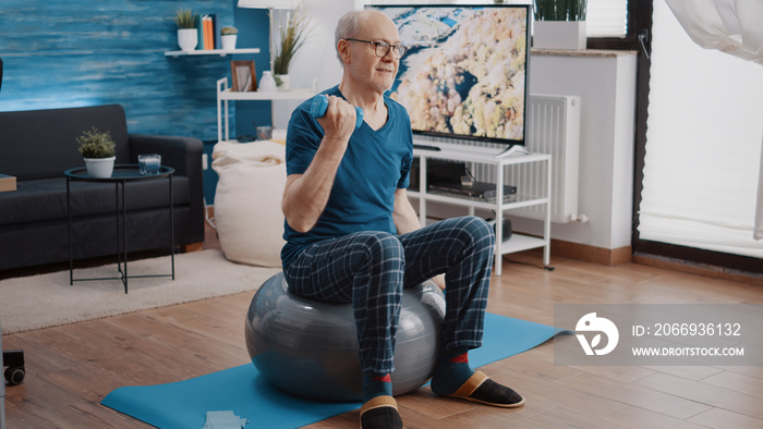 Senior man lifting dumbbells and sitting on fitness toning ball to do physical training and exercise. Retired person doing workout activity with weights to work arms muscles at home.