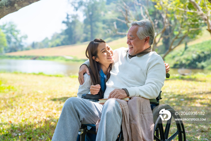 Happy Asian woman caring and hugging senior man grandfather sitting on wheelchair in the park. Elderly retired male relax and enjoy outdoor leisure activity with daughter. Family relationship concept.