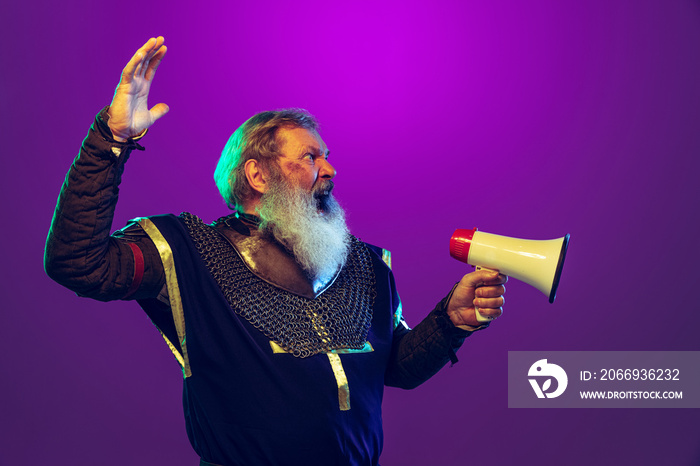 Funny old grey-bearded man in image of medieval warrior or knight in period costume shouting at megaphone isolated o purple background in neon. Comparison of eras