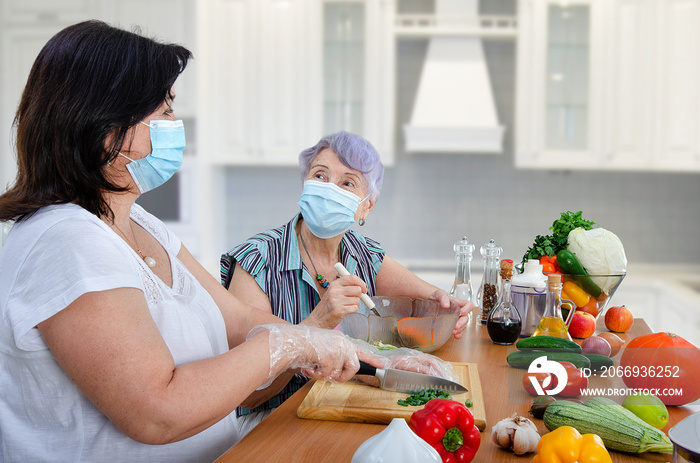 Despite the coronavirus pandemic, a caregiver and a senior adult woman cook together a vegetable salad once a week in the kitchen. Both put on protective masks.