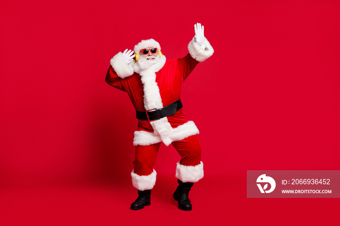 Full length photo of pensioner old man grey beard dancing carefree wear santa costume gloves coat belt sunglass headwear wireless earphones black leather boots isolated red color background