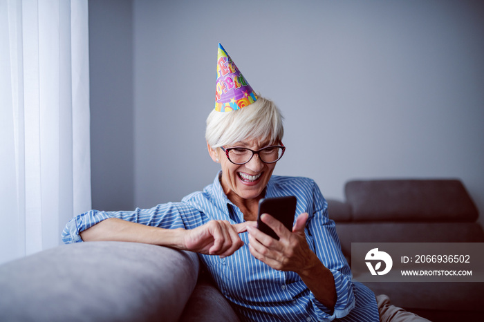 Cheerful caucasian senior woman with eyeglasses and with birthday cap on head sitting on sofa in living room next to window and reading birthday messages on smart phone.