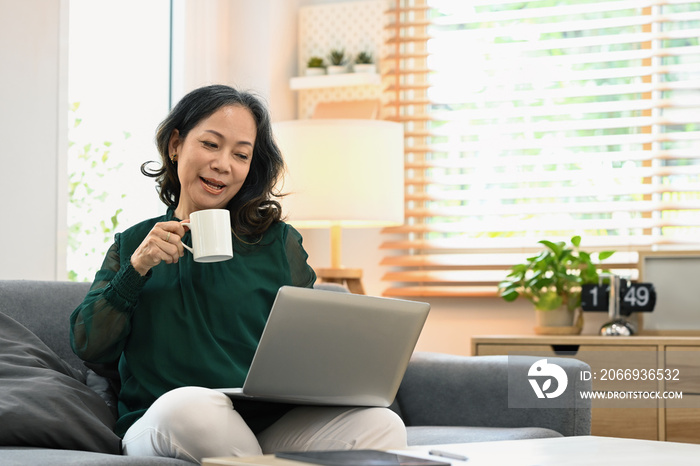 Joyful middle aged woman in casual clothes drinking coffee and using a laptop on couch at home