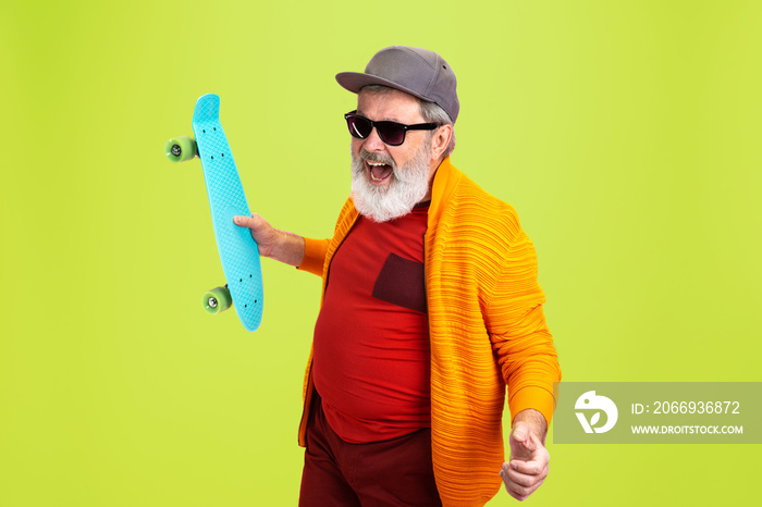 Skater man. Portrait of senior hipster man in fashionable eyewear isolated on green studio background. Tech and joyful elderly lifestyle concept. Trendy colors, forever youth. Copyspace for your ad.