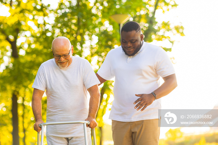 Caregiver is teaching old man to walk with walker. Professional nurse and patient walking outdoor at sunset. Assistance, rehabilitation and health care.