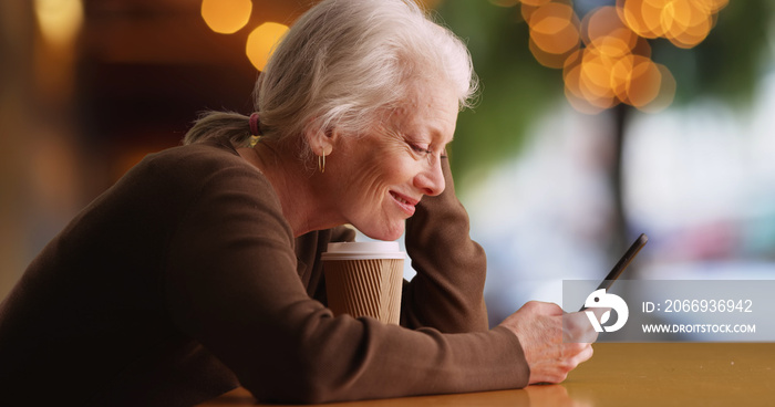 Happy elderly white woman using mobile device sitting outside at coffee shop