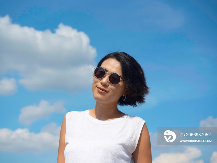 Happy woman in summertime. Smiling beautiful Asian woman short hair wearing sunglasses and casual white sleeveless shirt looking at camera on blue sky and cloud background on sunny day in summer.