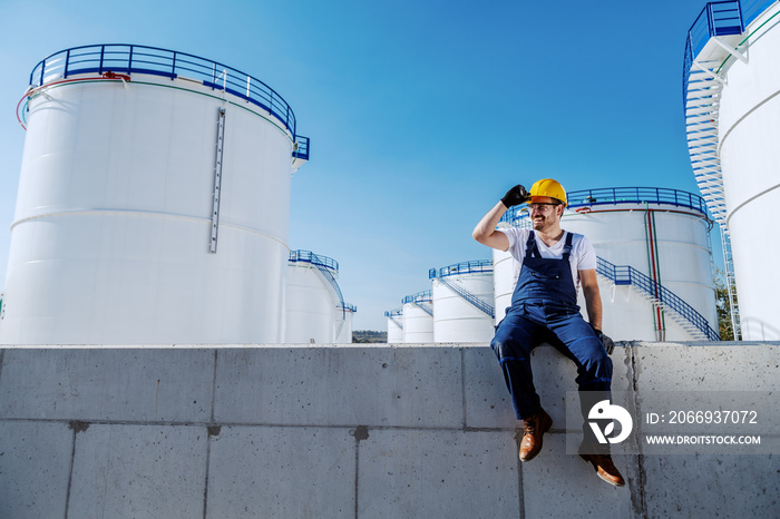 Full length of handsome caucasian workman in overalls and with helmet on head sitting on wall and looking away. In background are tanks with oil. Refinery exterior.