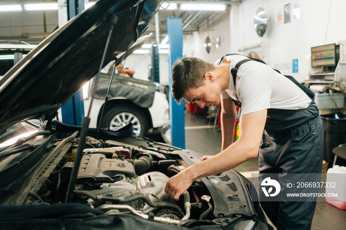 Side view of focused handsome professional male car mechanic in blue uniform standing in front of open hood, inspecting engine of car coming in for repair or maintenance in auto repair workshop..