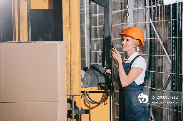 Smiling workwoman talking on walkie talkie while standing near forklift loader