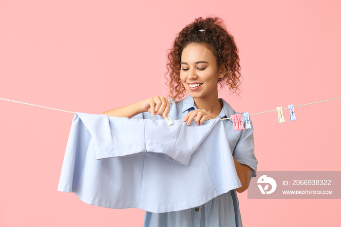 Young African-American woman hanging clean skirt with clothespin on pink background