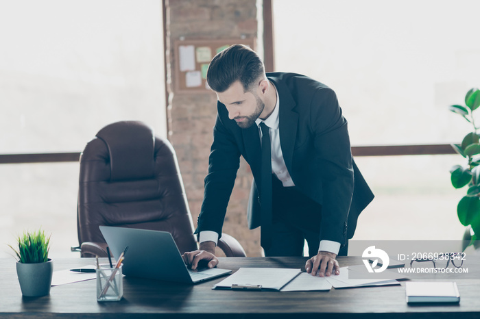 Photo of handsome successful business guy typing notebook keyboard check compare paper contract details online stats wear black blazer shirt tie suit leaning table office indoors