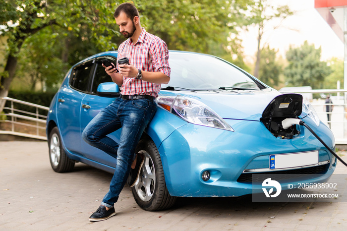 Young handsome man use phone and drink coffee while his electro car charging at station.