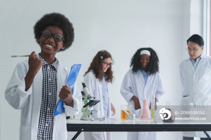 Little cute and smart African boy is taking notes about the science experiment in the laboratory. With his diversity African American Caucasian friends and Asian teacher doing experiment on background