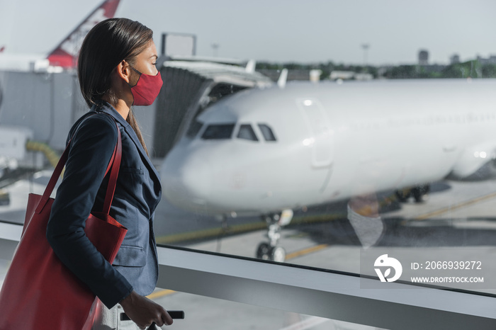 COVID-19 Travel by plane during coronavirus. Asian tourist waiting for flight at terminal wearing face mask indoors looking at window with airplane on tarmac.