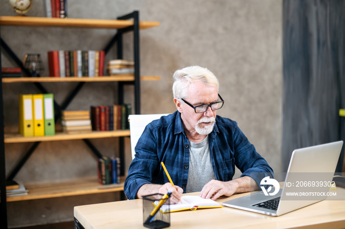 Mature man in casual wear and eyeglasses is using a laptop computer for online studying at home office. A gray-haired man is writing in notebook while sitting and watching webinars, online classes