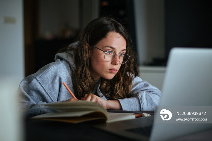 Young millennial woman at home look at laptop and make notes in notebook. Learn new skill from online classes or masterclass. New generation use technology for education and freelance