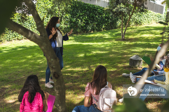 Shot from behind a tree of a Latina teacher teaching with a mask in the open air.