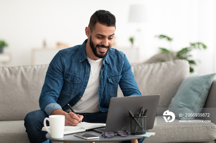 Happy young Arab guy working or studying on laptop computer, taking notes, having business meeting indoors