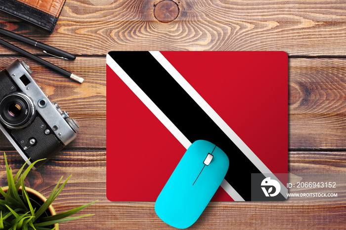 Trinidad And Tobago flag on wooden background with blue wireless mouse on a mouse pad, top view. Digital media concept.