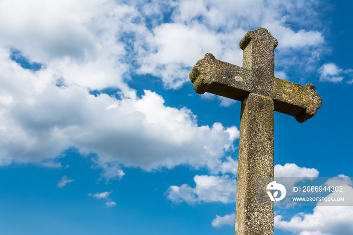 Old gothic Christian stone cross on dramatic blue sky background with white clouds. Detail of sunlit ancient crucifix. Vintage monument with moss and lichen cover. Idea a death or god, faith and hope.