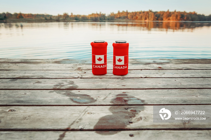 Two cans of beer in red cozy beer can coolers with Canadian flag standing on wooden pier by lake outdoor. Wet footprints on wooden dock. Friends celebrating Canada Day national celebration by water.