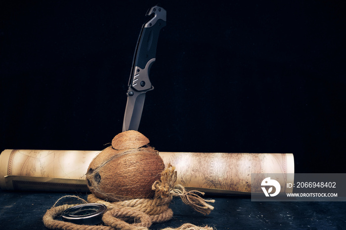 Tactical folding knife is stuck in split coconut lying next to rope and an old map on dark background.Survival and adventure.