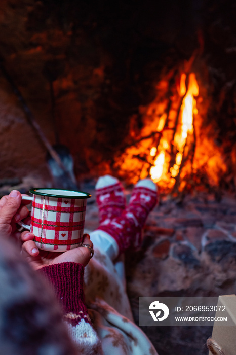 Close up of people holding christmas mug cup and drinking in front of a warm fireplace at home in chalet for december holiday season vacation. Female drink coffee and relax indoor leisure activity