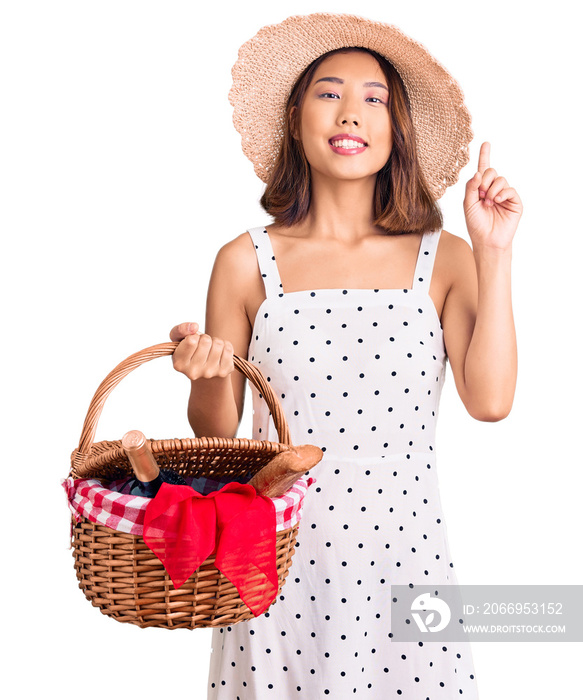 Young beautiful chinese girl wearing summer hat holding picnic basket surprised with an idea or question pointing finger with happy face, number one