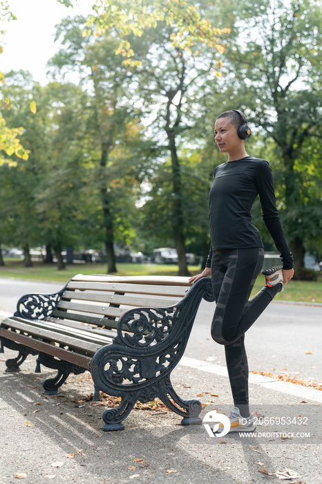 Woman in headphones and sports clothing stretching leg in park