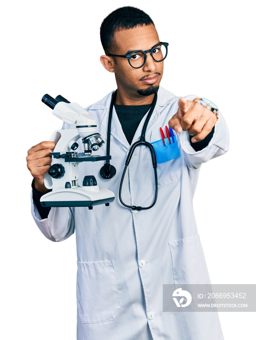 Young african american man wearing scientist uniform holding microscope pointing with finger to the camera and to you, confident gesture looking serious