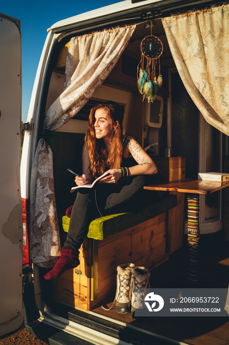 Chica pelirroja en una furgoneta camper camperizada en una zona costera de playa al atardecer leyendo y escribiendo con estilo libre hippie