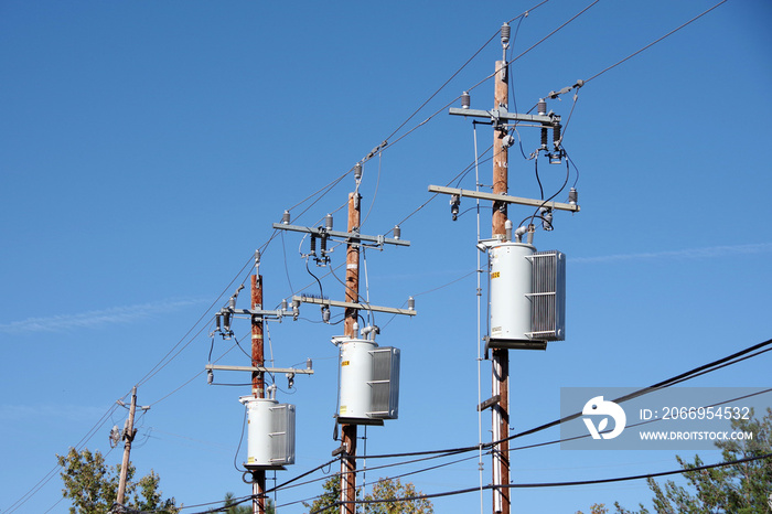 Low angle upward view of a row of tall electricity distribution poles with transformers and power lines seen against a clear blue sky