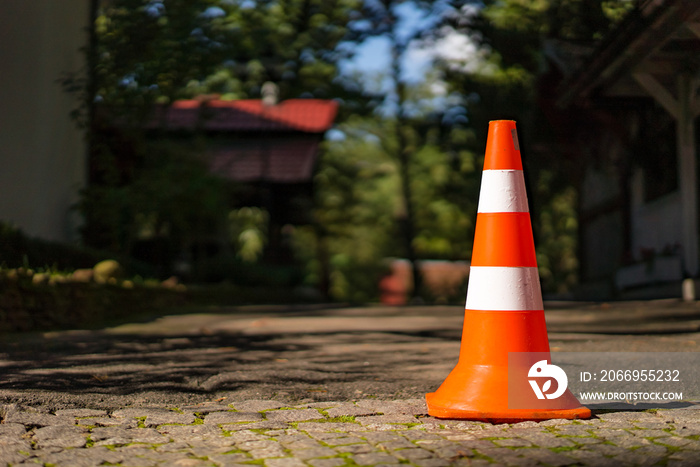 Red orange cone with a white stripe on the paving stone road. Drive safety and constructions concept. Closeup. Blurred background with green trees and buildings. Sunlight and shadow.