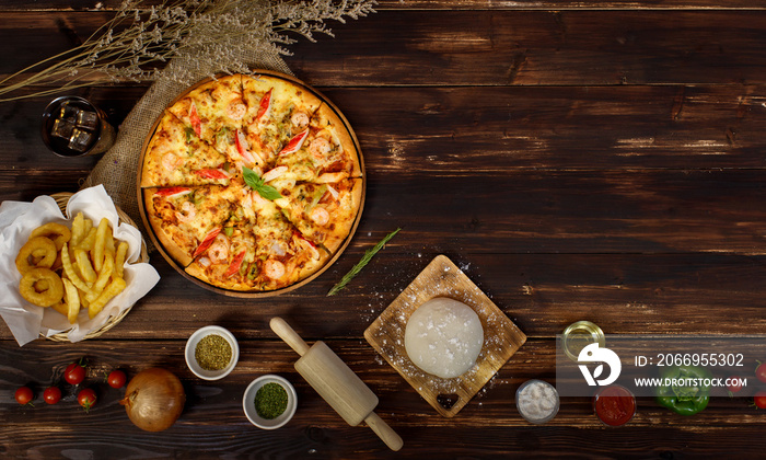 Tray of homemade seafood pizza surrounded by ingredients located in row below the wooden background and decorated with dried plants, cold beverage and fried potatoes with copy space.