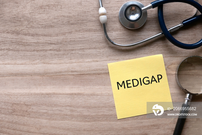 Top view of stethoscope, magnifying glass and yellow paper note written with Medigap on wooden background with copy space.