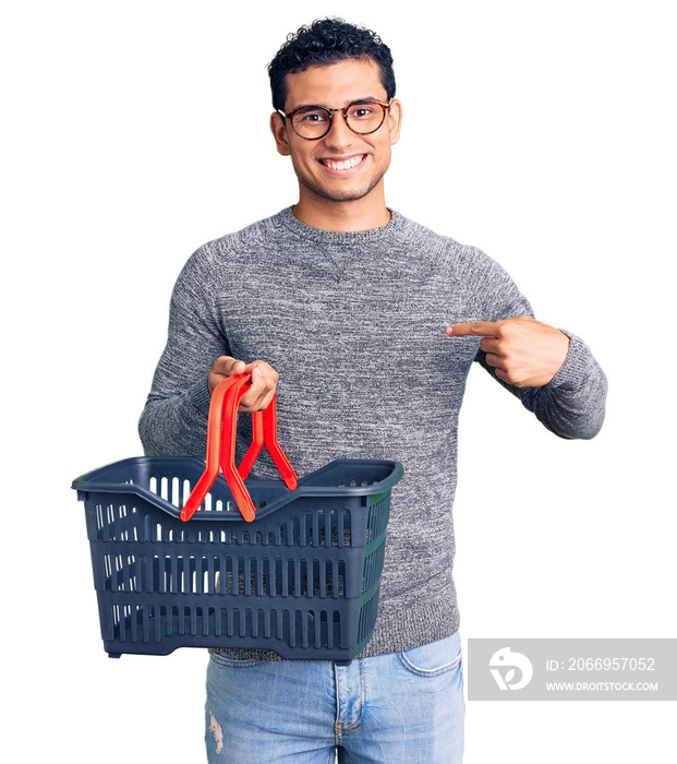 Hispanic handsome young man holding supermarket shopping basket pointing finger to one self smiling happy and proud