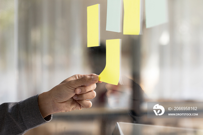 Note paper pasted on glass in meeting room, businessman’s hand pointing and pasting note paper to write messages or meeting topics. The idea of writing a notepad to memorize.