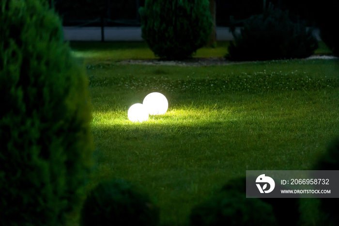 a pair of white ball-shaped lighting fixtures on a green lawn illuminate the landscape of a spring evening park with thuja bushes, the glow of ground lanterns, nobody.