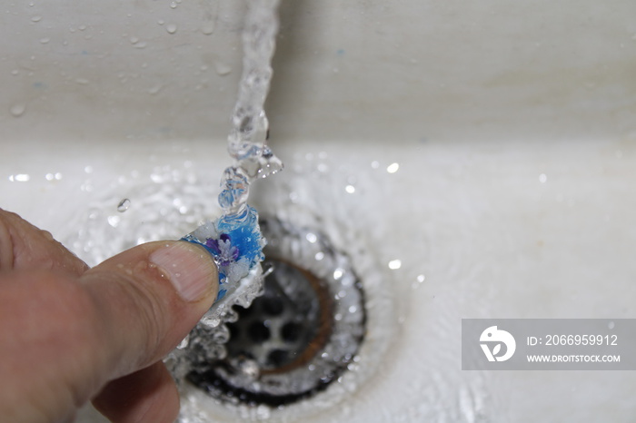 Lava a mano el cepillo de dientes en el baño de la casa para la limpieza oral con cerdas de plástico bajo agua corriente, forma un diseño original de higiene personal con fondo de la pileta blanca