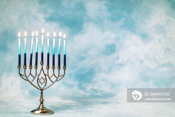 A silver menorah for the Jewish holiday Hanukkah with burning glowing eight candles on fire lit up on a blue background with copy space