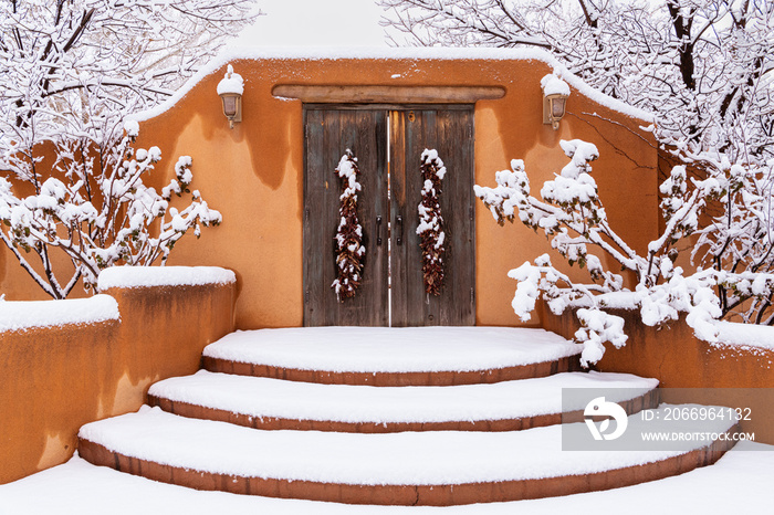 Winter scene of snow-covered adobe wall with rustic wood doors and chile ristras in Santa Fe, New Mexico