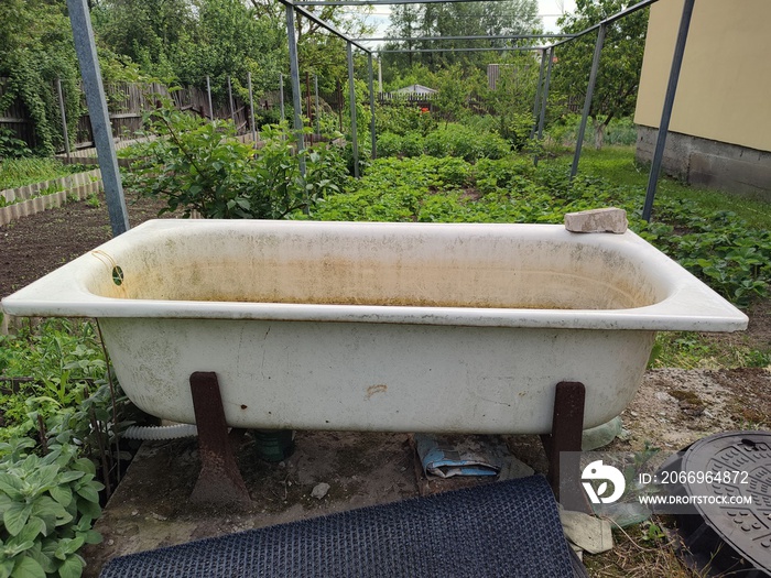 n old dirty enameled bathtub stands on rusty iron supports in a green garden in summer