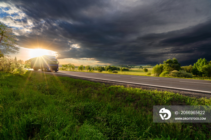 Blue truck driving on the asphalt road in rural landscape in the rays of the sunset with dark storm cloud