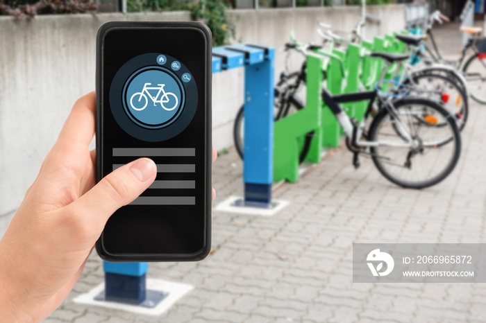 technology, transport and sustainability concept - close up of hand holding smartphone with mobile app and bicycle icon on screen over electric bike parking and charging station in city on background