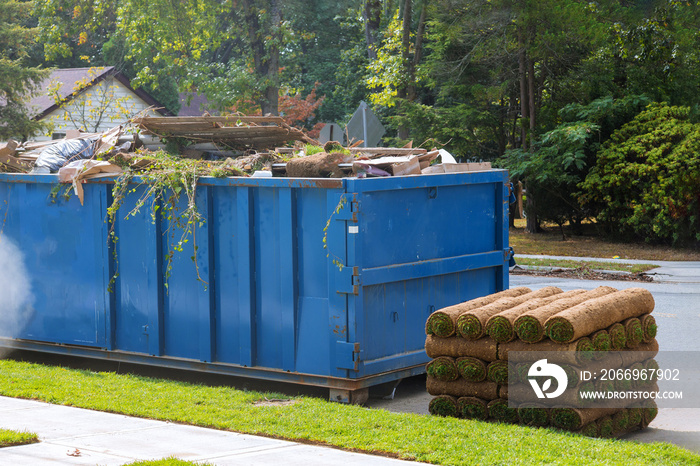 Lawn grass in rolls on pallets against of the street the rolled grass lawn is ready for laying and loaded dumpster near a construction