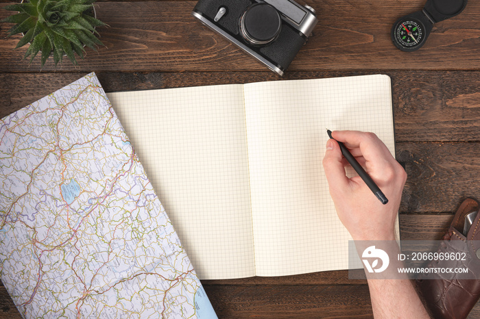 Travel planning concept. Hand writing in blank notepad with compass, knife, vintage camera and map on wooden table background.